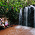 A family of five sits on a rock by a small waterfall in a lush, green forest, watching the water flow into a shallow reddish-brown pool.