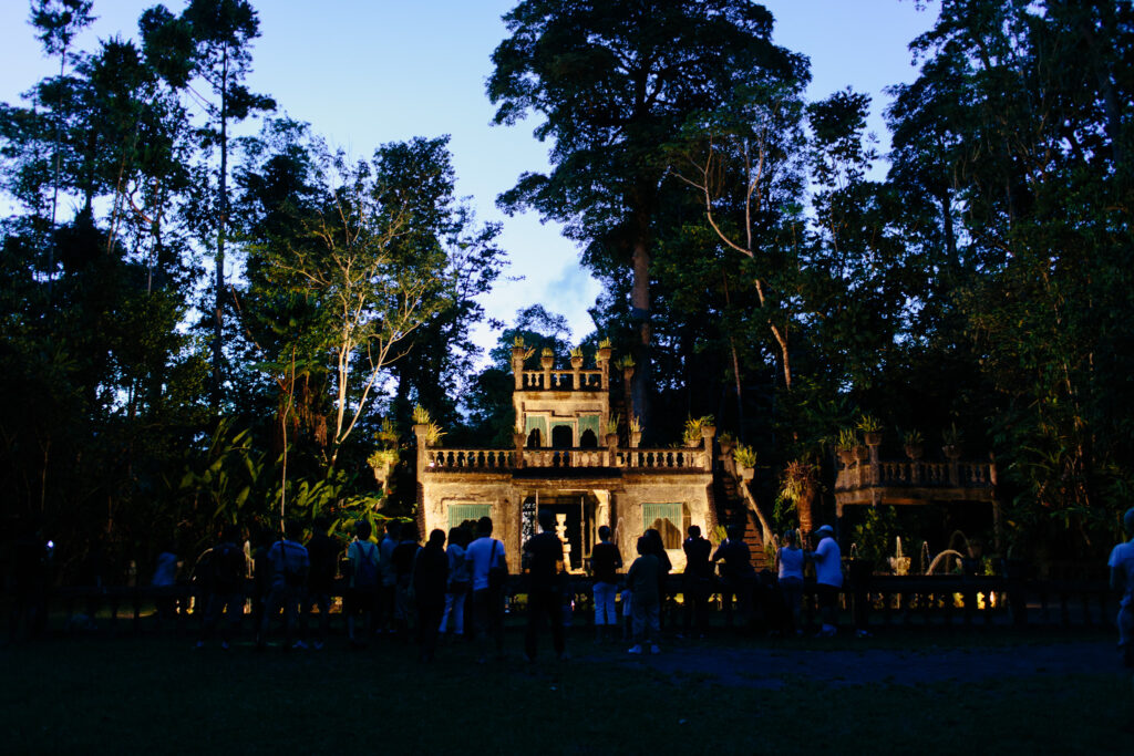 A group of people stands in front of a historic, castle-like structure surrounded by dense trees at dusk.