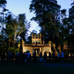 A group of people stands in front of a historic, castle-like structure surrounded by dense trees at dusk.