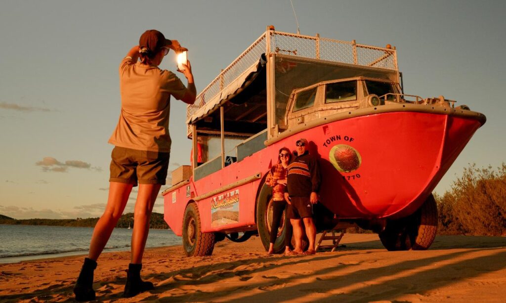 Person taking a photo of three people posing by a red amphibious vehicle on a sandy beach at sunset.