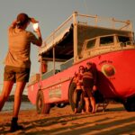 Person taking a photo of three people posing by a red amphibious vehicle on a sandy beach at sunset.