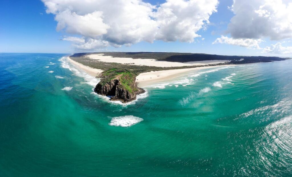 Aerial view of a coastal landscape with turquoise water, sandy beaches, rocky headland, and low clouds in a blue sky.
