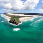 Aerial view of a coastal landscape with turquoise water, sandy beaches, rocky headland, and low clouds in a blue sky.