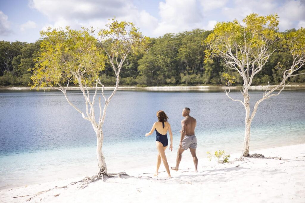 Two people in swimsuits walk along a sandy beach between trees beside a calm body of water, with a forested area in the background.