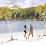 Two people in swimsuits walk along a sandy beach between trees beside a calm body of water, with a forested area in the background.