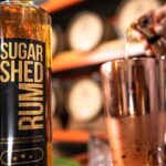 A close-up of a Sugar Shed Rum bottle beside a copper shaker, with a hand pouring liquid into the shaker; barrels are blurred in the background.