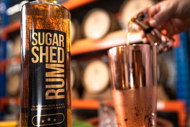 A close-up of a Sugar Shed Rum bottle beside a copper shaker, with a hand pouring liquid into the shaker; barrels are blurred in the background.