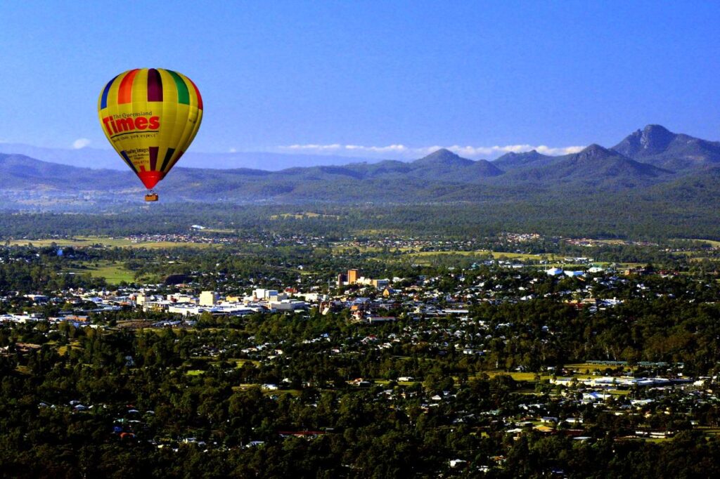 A colorful hot air balloon floats above a town surrounded by trees, with mountains visible in the background under a clear blue sky.
