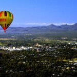 A colorful hot air balloon floats above a town surrounded by trees, with mountains visible in the background under a clear blue sky.