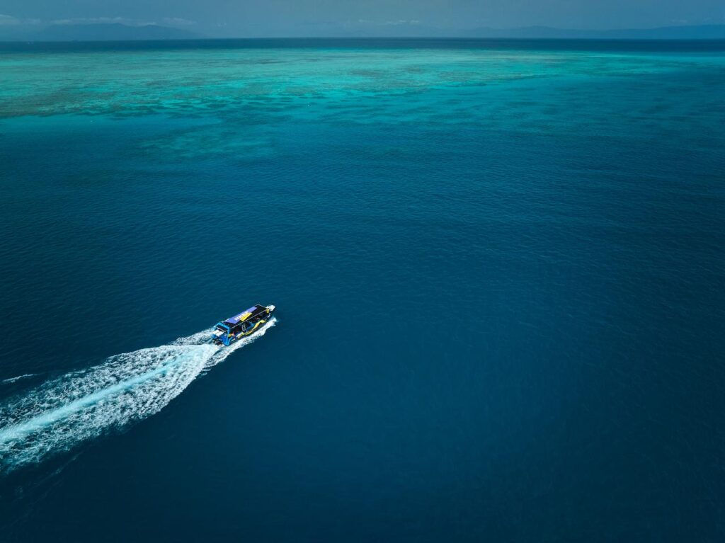 A single speedboat moves across deep blue ocean water, leaving a white wake, with shallow turquoise reef visible in the distance.