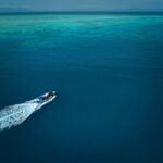 A single speedboat moves across deep blue ocean water, leaving a white wake, with shallow turquoise reef visible in the distance.