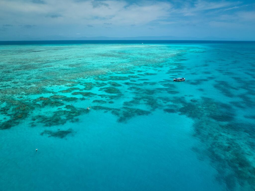 Aerial view of a boat floating on clear turquoise water above coral reefs under a partly cloudy sky.
