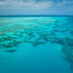 Aerial view of a boat floating on clear turquoise water above coral reefs under a partly cloudy sky.