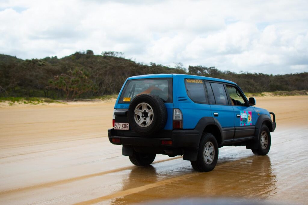 A blue SUV with a spare tire drives on a sandy beach with trees and hills in the background under a cloudy sky.