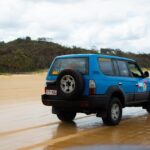 A blue SUV with a spare tire drives on a sandy beach with trees and hills in the background under a cloudy sky.