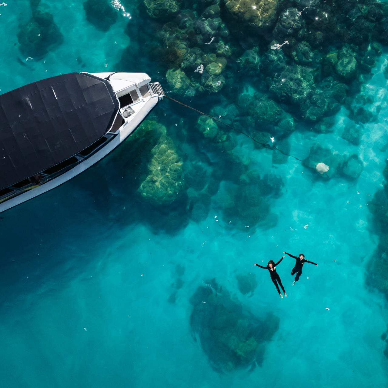 Aerial view of two people snorkeling in clear turquoise water near a boat anchored above a rocky seabed.
