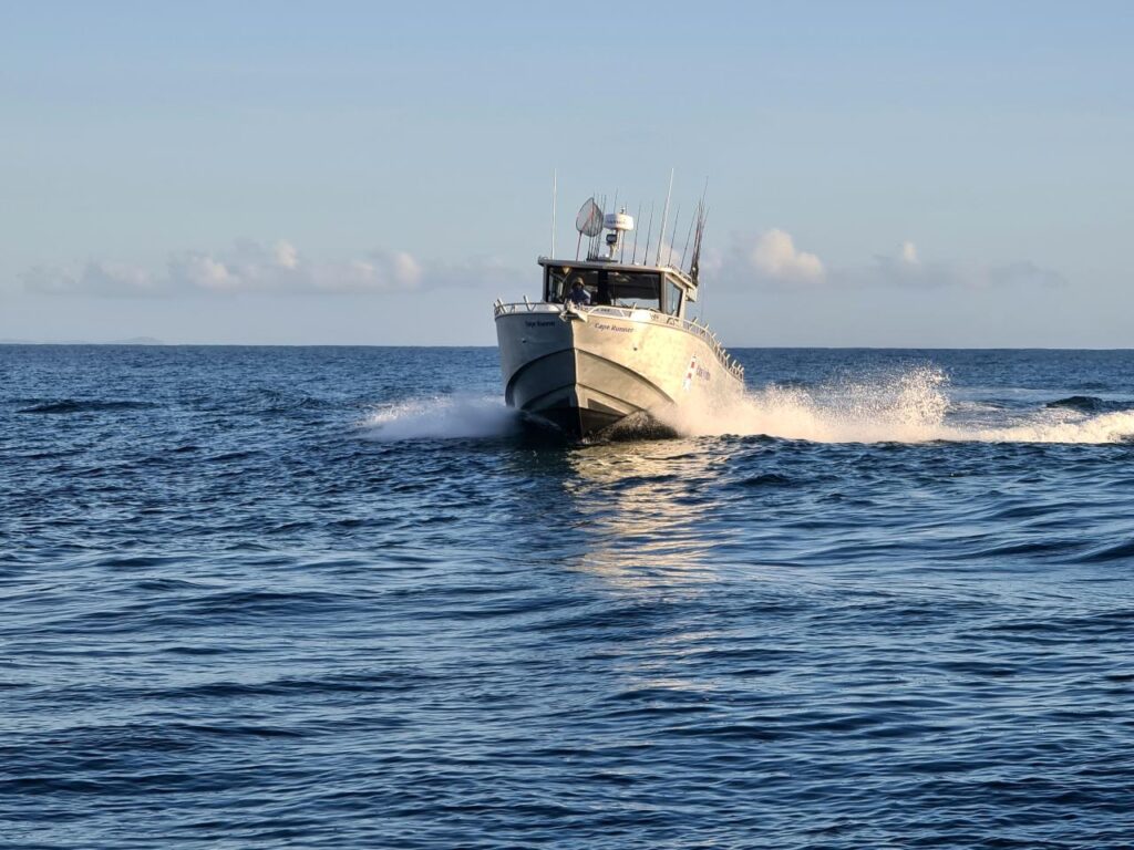 A white motorboat speeds across calm, blue ocean water under a clear sky, creating a trail of waves behind it.