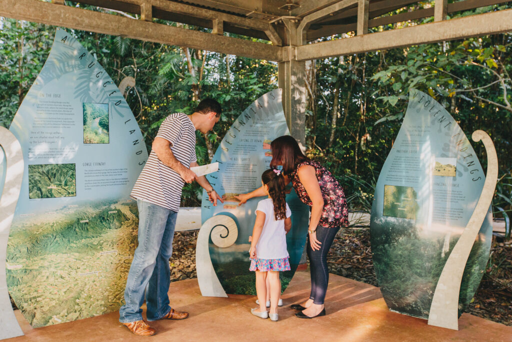 Three people, two adults and a child, read and point at interpretive display panels under a shelter in an outdoor nature area.
