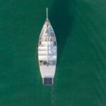 Aerial view of a white sailboat with a solar panel on its deck, docked in calm green water near other boats.