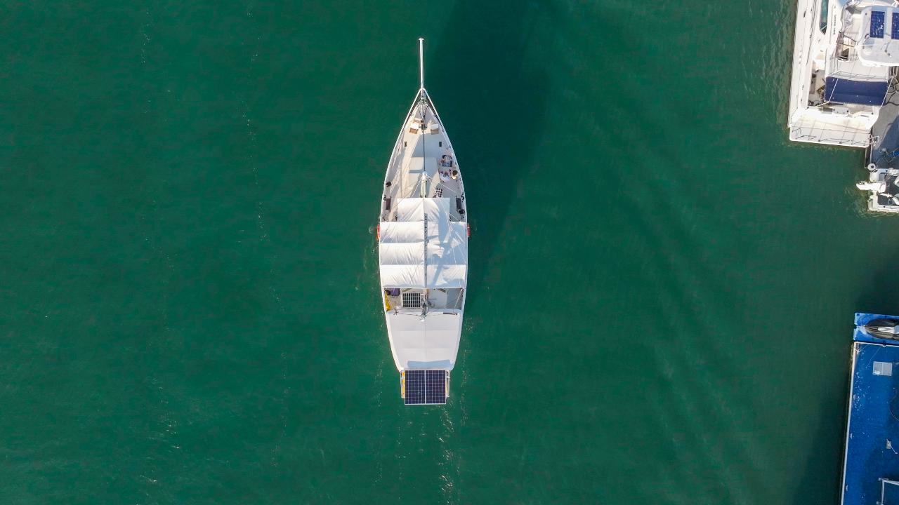 Aerial view of a white sailboat with a solar panel on its deck, docked in calm green water near other boats.