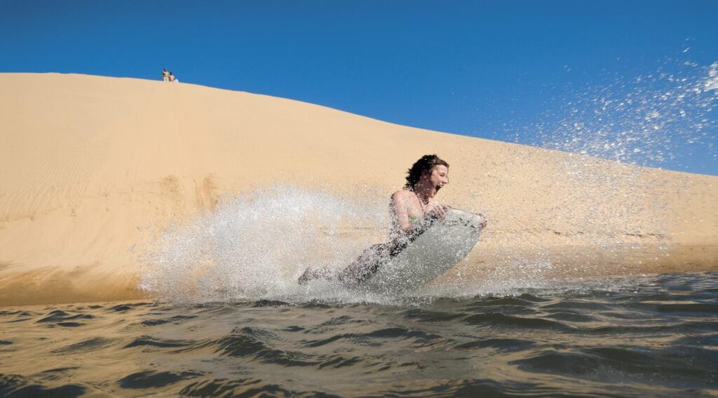 Person riding a sandboard down a steep sand dune into water, creating a large splash, with a clear blue sky above.