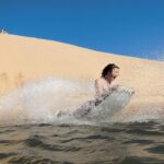 Person riding a sandboard down a steep sand dune into water, creating a large splash, with a clear blue sky above.