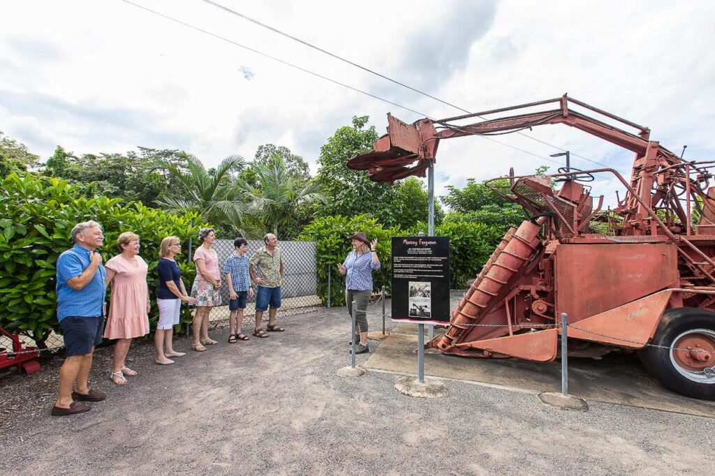 A group of people listens to a guide explaining an old, red agricultural machine displayed outdoors, with greenery and a partly cloudy sky in the background.