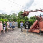A group of people listens to a guide explaining an old, red agricultural machine displayed outdoors, with greenery and a partly cloudy sky in the background.