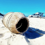 A large, rusted barrel lies on white sand near the shore, with a parked vehicle and camper in the background under a clear blue sky.