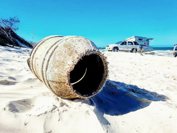 A large, rusted barrel lies on white sand near the shore, with a parked vehicle and camper in the background under a clear blue sky.