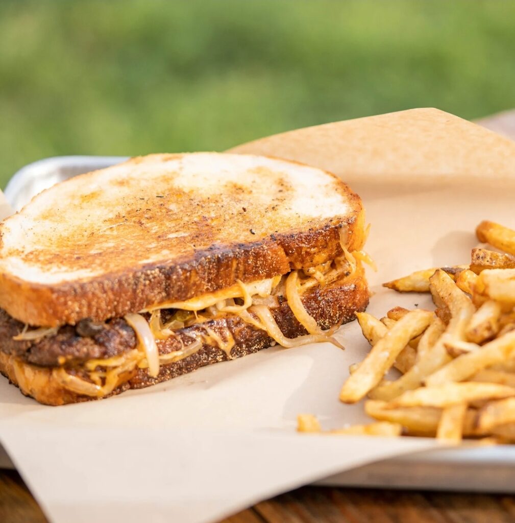 A grilled sandwich filled with beef and sautéed onions is served on parchment paper next to a pile of French fries.