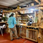 A man stands at a rustic bar counter while a bartender points at a menu above; shelves hold bottles and decor, and the setting features wood and corrugated metal.