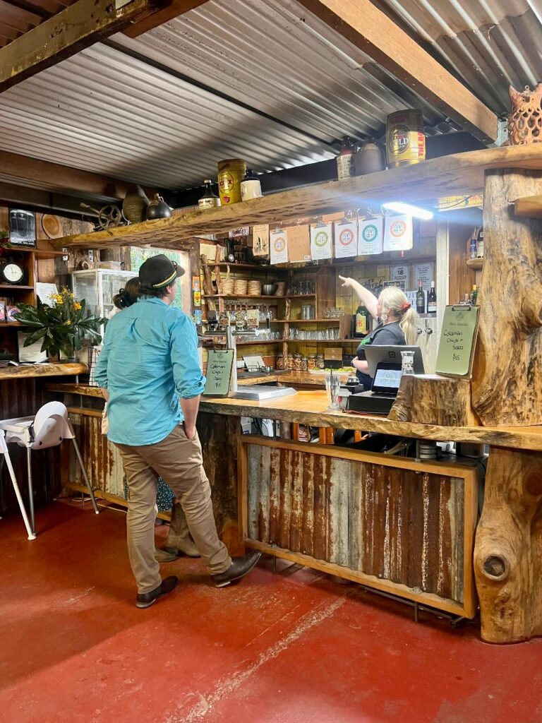 A man stands at a rustic bar counter while a bartender points at a menu above; shelves hold bottles and decor, and the setting features wood and corrugated metal.