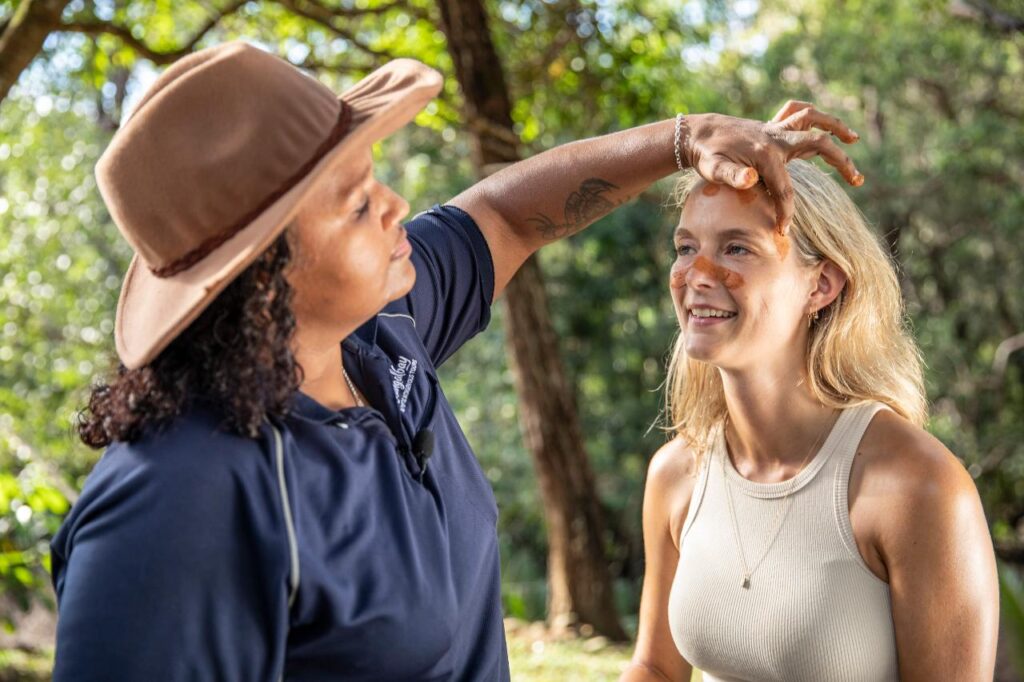 A woman applies ochre to another woman's face outdoors, both smiling, with trees and sunlight in the background.