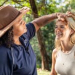 A woman applies ochre to another woman's face outdoors, both smiling, with trees and sunlight in the background.