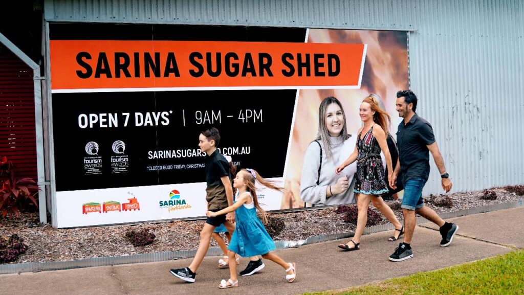 A family of four walks on a sidewalk past a large sign for Sarina Sugar Shed, which advertises its opening hours and website.