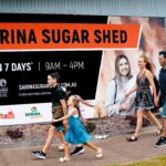 A family of four walks on a sidewalk past a large sign for Sarina Sugar Shed, which advertises its opening hours and website.