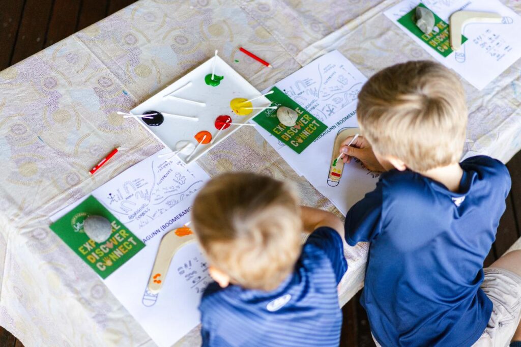 Two children sit at a table painting on paper with colored paints and brushes. Art supplies and painting guides are laid out in front of them.