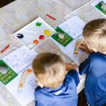 Two children sit at a table painting on paper with colored paints and brushes. Art supplies and painting guides are laid out in front of them.