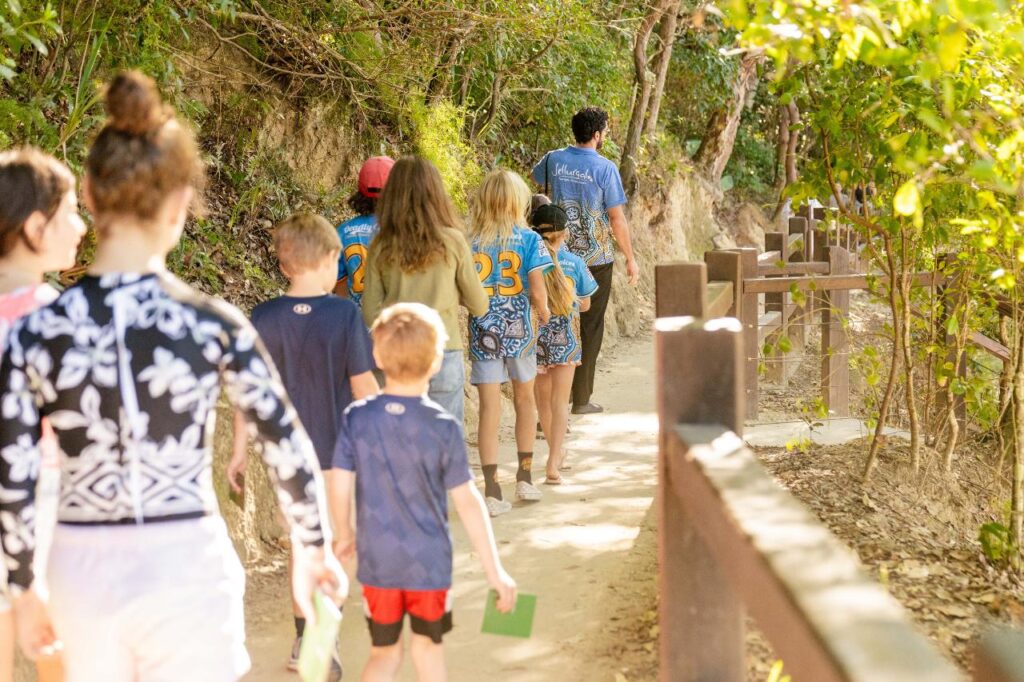 A group of children and adults walk along a dirt path surrounded by trees, with some holding green cards and wearing matching shirts with numbers.