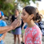 A person applies white paint to a girl's face during an outdoor group activity near a rocky shoreline. Other people stand in the background.