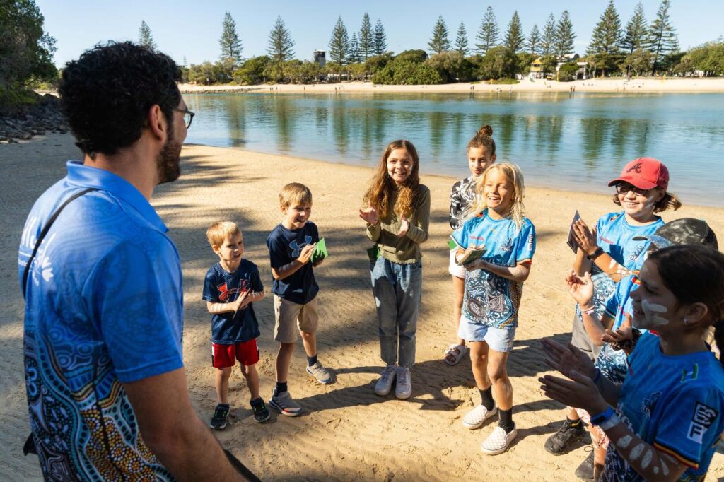 A group of children and an adult stand on a sandy lakeshore, smiling and clapping, with water and trees in the background.