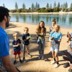 A group of children and an adult stand on a sandy lakeshore, smiling and clapping, with water and trees in the background.