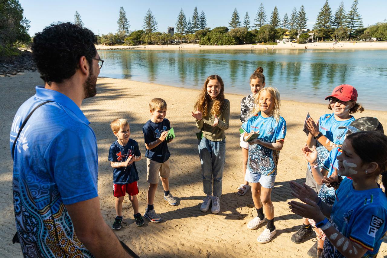 A group of children and an adult stand on a sandy lakeshore, smiling and clapping, with water and trees in the background.