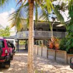A stilt house with a wraparound porch is surrounded by tropical plants, with two magenta vehicles parked in the sandy driveway under palm trees.