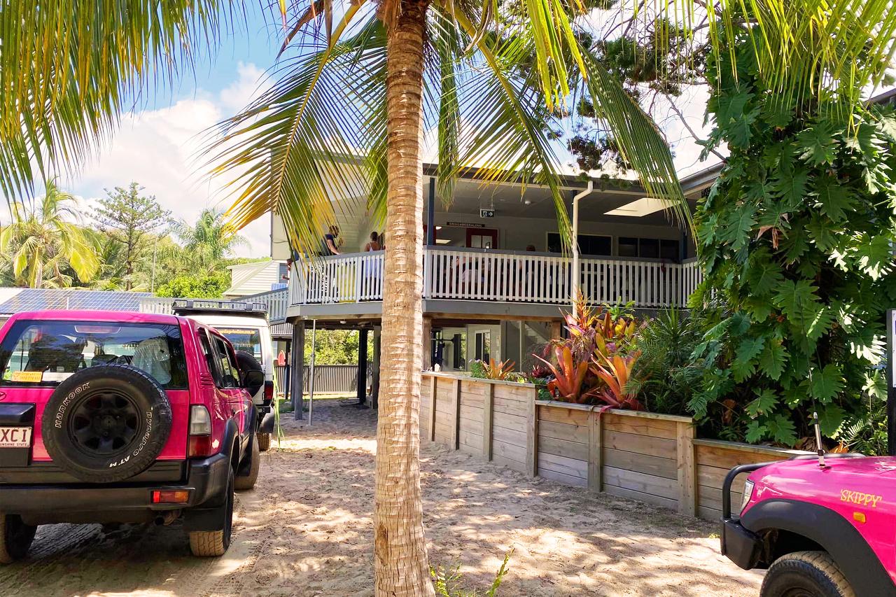 A stilt house with a wraparound porch is surrounded by tropical plants, with two magenta vehicles parked in the sandy driveway under palm trees.