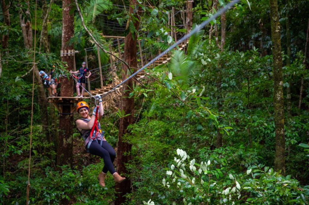 A person wearing safety gear is ziplining through a lush green forest, with others waiting on a platform in the background.