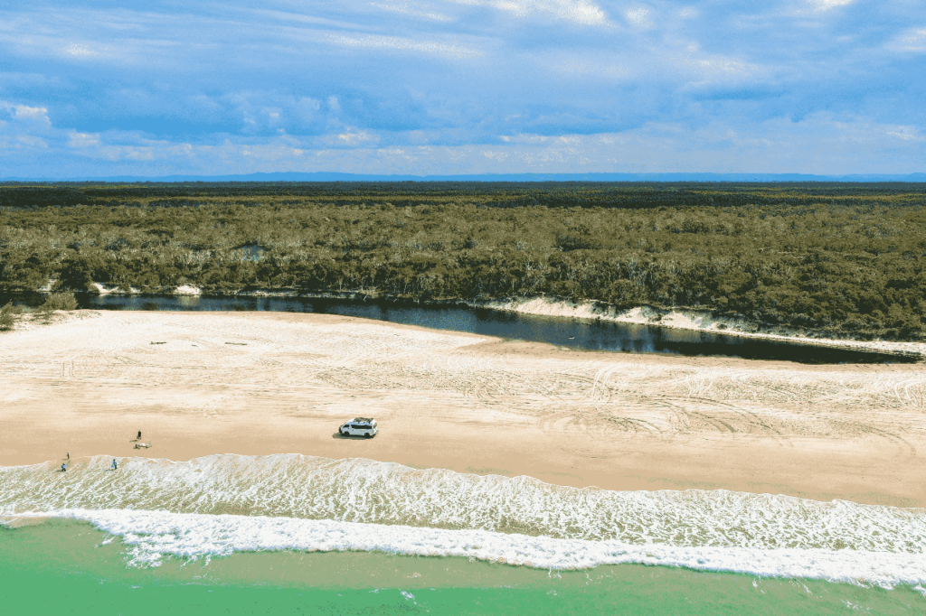 Aerial view of a sandy beach with a parked vehicle near the shoreline, a river running parallel to the sea, and a forest in the background under a partly cloudy sky.