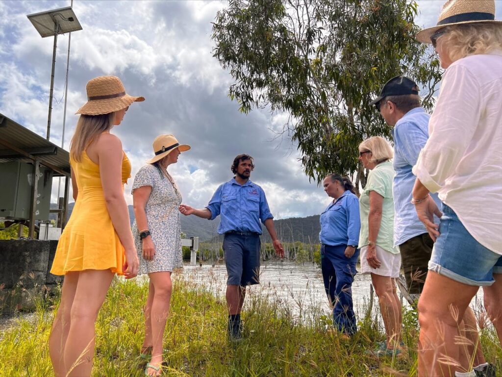 A group of people, some wearing hats, stand and listen to a man in a blue shirt speaking near a body of water and grass under a cloudy sky.