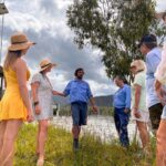 A group of people, some wearing hats, stand and listen to a man in a blue shirt speaking near a body of water and grass under a cloudy sky.